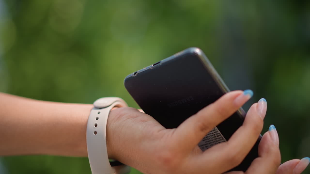Commute closeup caucasian woman hands with smartphone connecting wired headphones in shaded park, white cable threading over fitness wristband, focused fingers with neat manicure, soft bokeh, short