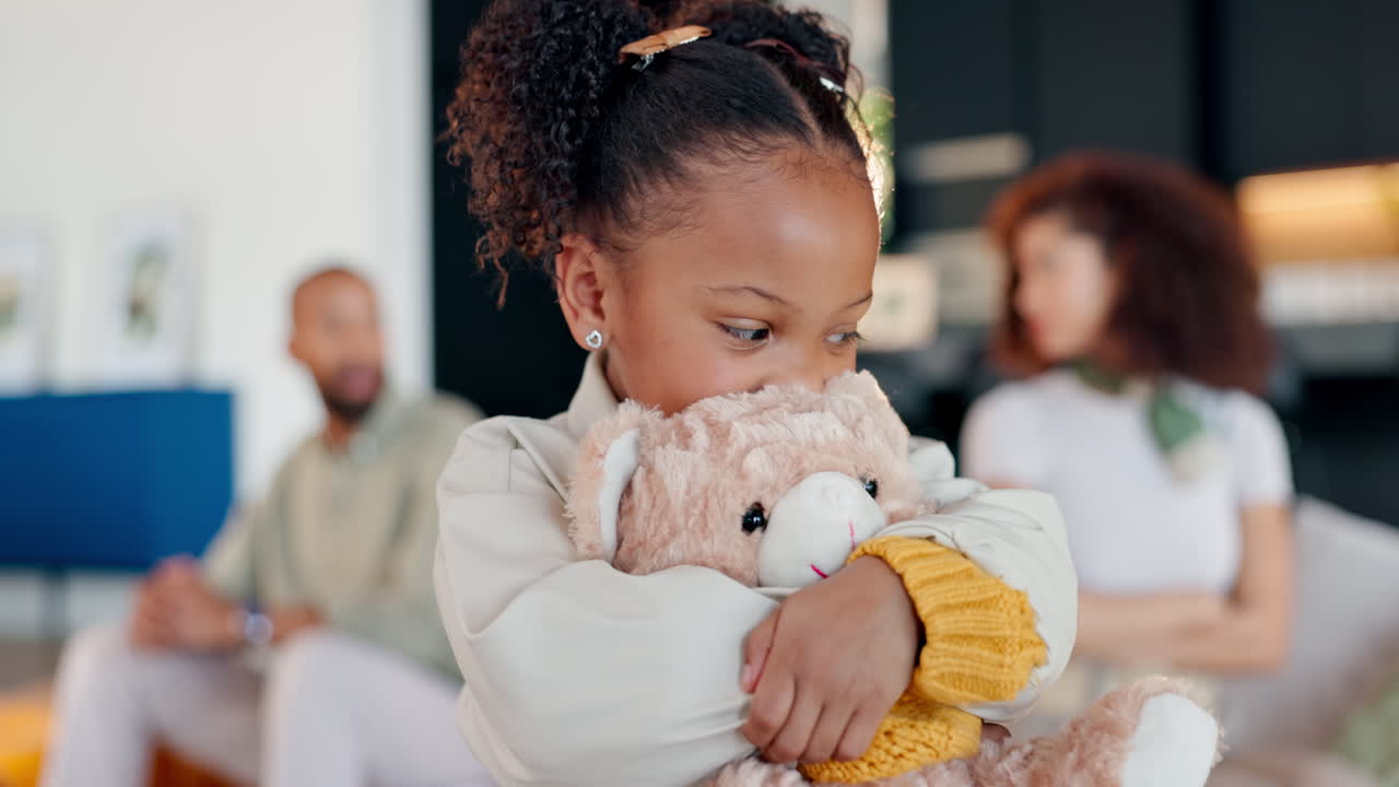 A young girl hugs her teddy bear while her parents sit in the background
