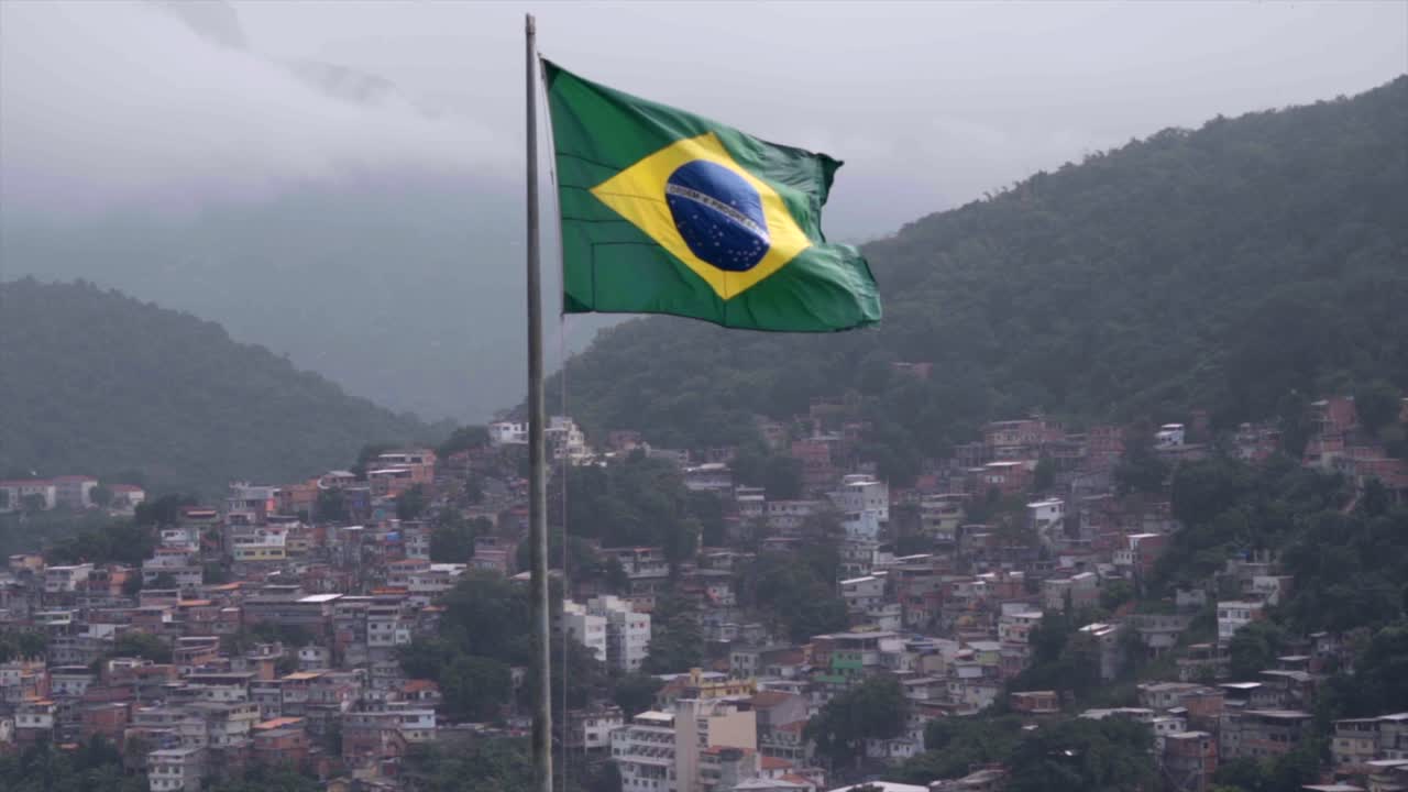 Gran bandera brasileña ondeando al viento frente a una gran favela en Copacabana, con colinas neblinosas y nubladas visibles al fondo. Toma estática en cámara lenta.