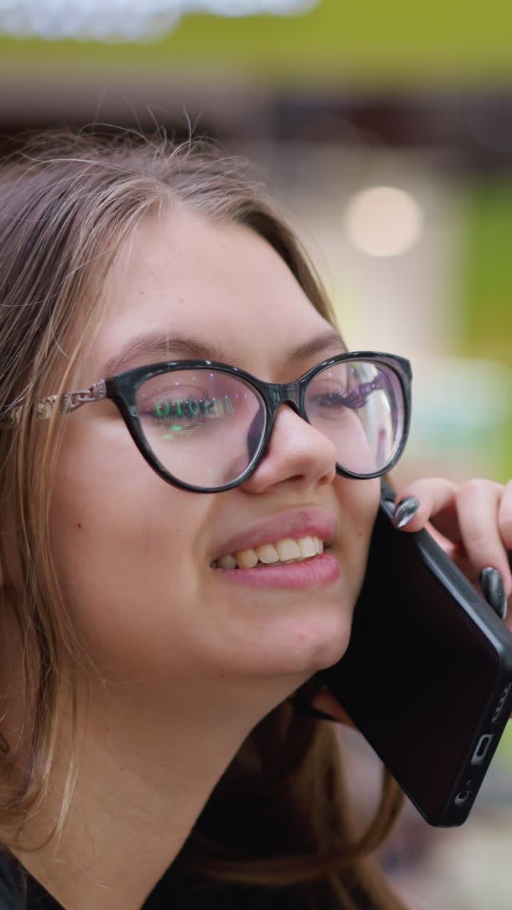 primer plano de una mujer alegre hablando por teléfono con camiseta negra y gafas, con fondo borroso con efecto de luz bokeh y estructuras modernas