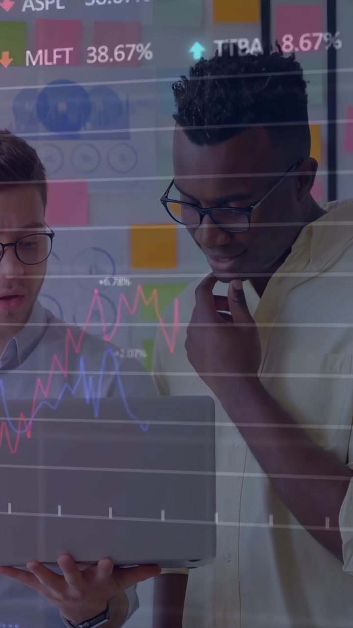Vertical video: Two men holding laptop, pointing, sparking finance chart overlay, analyzing trends