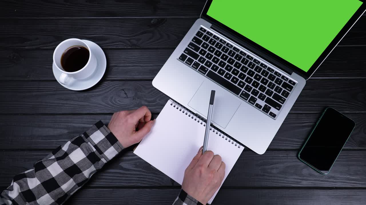 The girl got ready to write note from the laptop into a spiral notebook. Top view of the hands of a girl who sits at a table with a laptop with a green screen and a notebook. Slow motion. Close up
