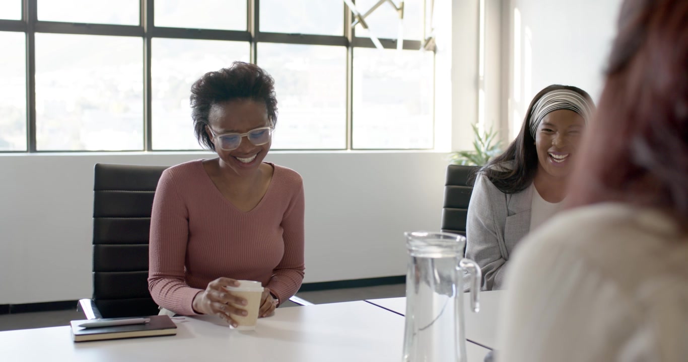 Diverse female coworkers smiling and listening in meeting room after speaker gesture holding coffee