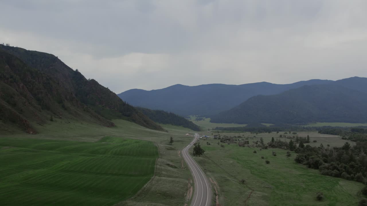 Riprese con drone di una strada asfaltata tortuosa che attraversa campi verdi e un'ampia valle di montagna sotto un cielo nuvoloso nell'Altai, Russia, un paesaggio rurale panoramico