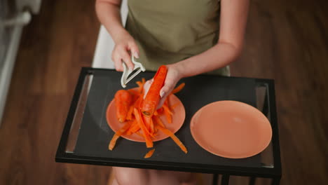 aerial view of chef in green shirt using handheld peeler to peel fresh carrot tray, carrot skins scattered on orange plate with empty plate beside, wooden floor and indoor lighting in kitchen