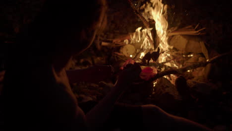 slow motion silhouette of man holding skewer of food over open campfire to cook it