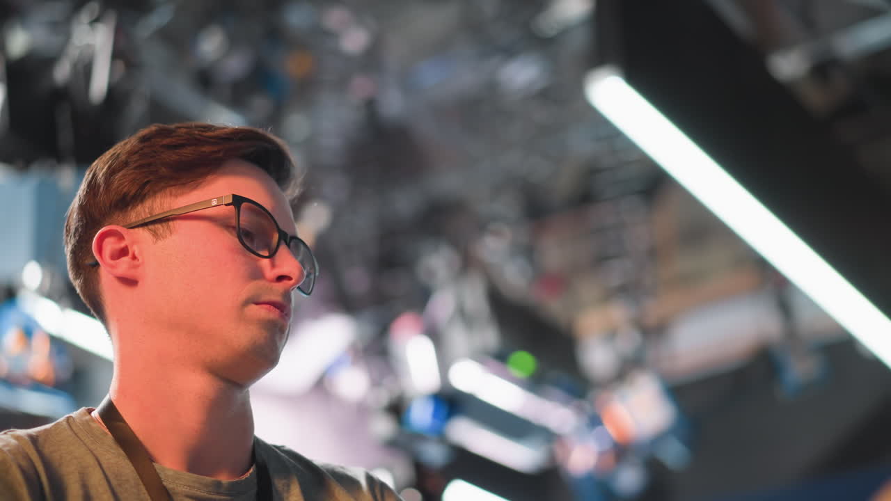 Professional person working in studio setup with lighting and equipment. Profile view shows focused individual adjusting system. Camera, lights, and modern studio technology in background
