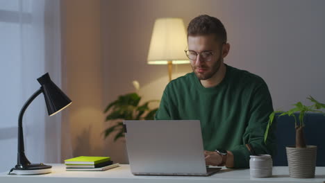 young man with glasses is typing on laptop at evening at home chatting on internet site medium portrait of intelligent guy