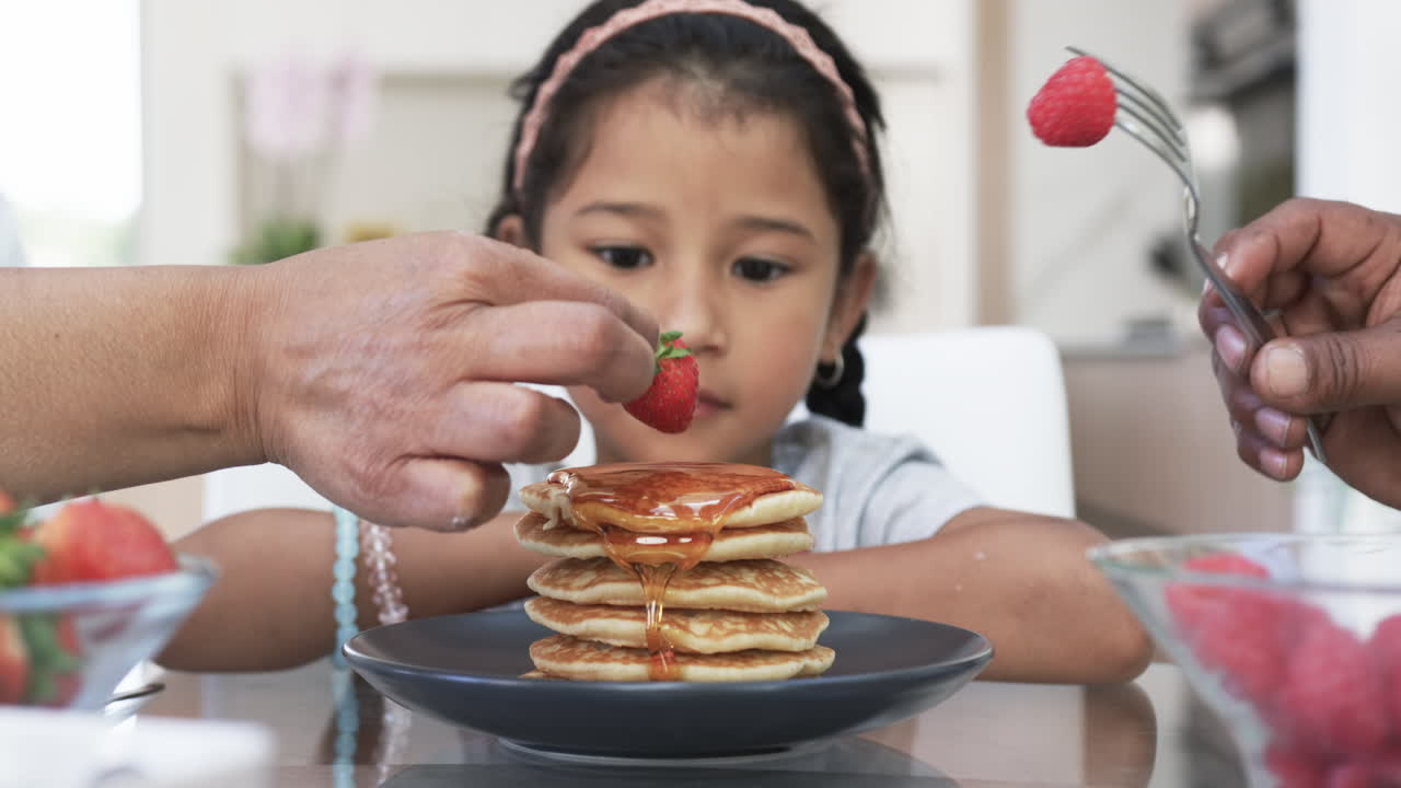 una chica biracial disfruta del desayuno mientras las manos la ayudan con jarabe y fruta.