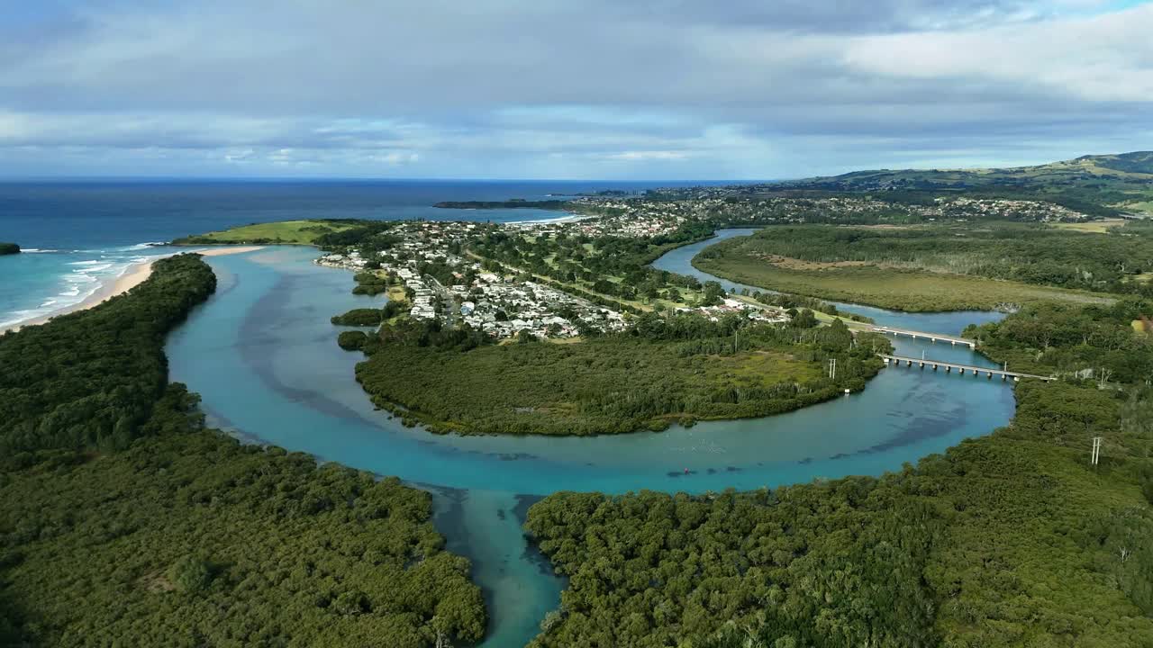 Drone gradual approach of winding Minnamurra River passing through estuary and wetlands under blue skies, to homes and development