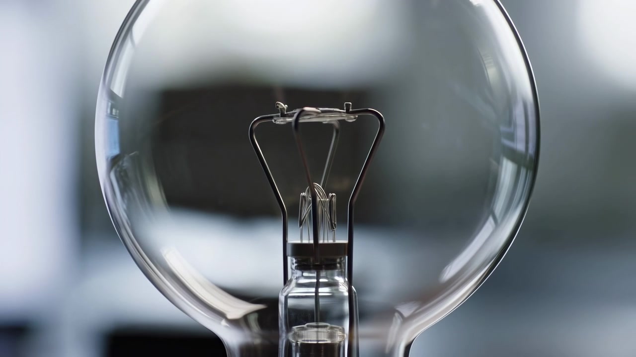 Symmetrical close-up of clear incandescent bulb held at eye level showing internal filament and support wires becoming visible through smooth glass globe against soft out-of-focus interior