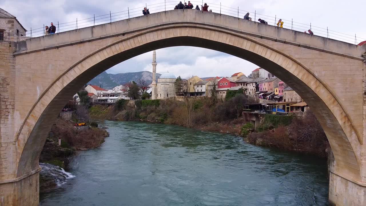 un dron con cámara vuela a lo largo del río neretva bajo un viejo puente peatonal arqueado en mostar, bosnia y herzegovina. los turistas caminan en el fondo de un hermoso paisaje urbano