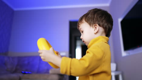dark-haired caucasian toddler in yellow sweater turns a ring from pyramid in hands. side view. baby boy having fun at home. low angle view.