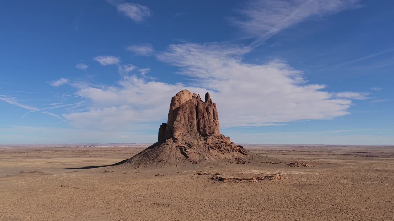 En droneflyvning bevæger sig mod en isoleret sandstensklippe (butte-monolit) i en stor, tør ørkenvildmark med barske geologiske formationer under en klar, blå himmel.
