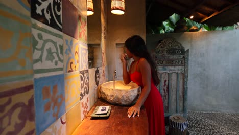 young tan asian woman putting on lipstick and makeup in traditional outdoor balinese bathroom with mosaic tile