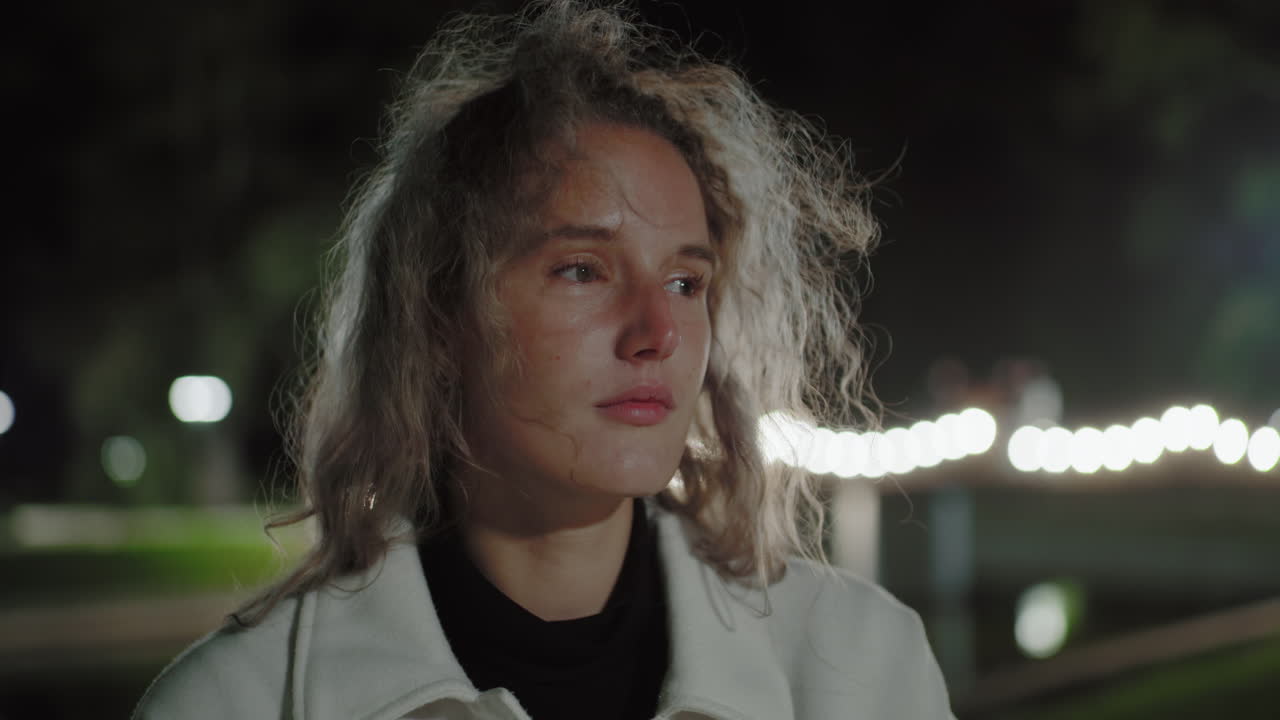 Nighttime scene of elegant student with curly hair fluttering in gentle breeze, alone in quiet setting, eyes momentarily closed then opened, shimmering lights reflecting softly
