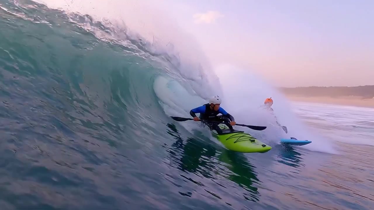 Due kayakisti cavalcano le onde al tramonto, navigando abilmente grandi onde oceaniche in un bellissimo paesaggio costiero con colori tenui e acque calme.