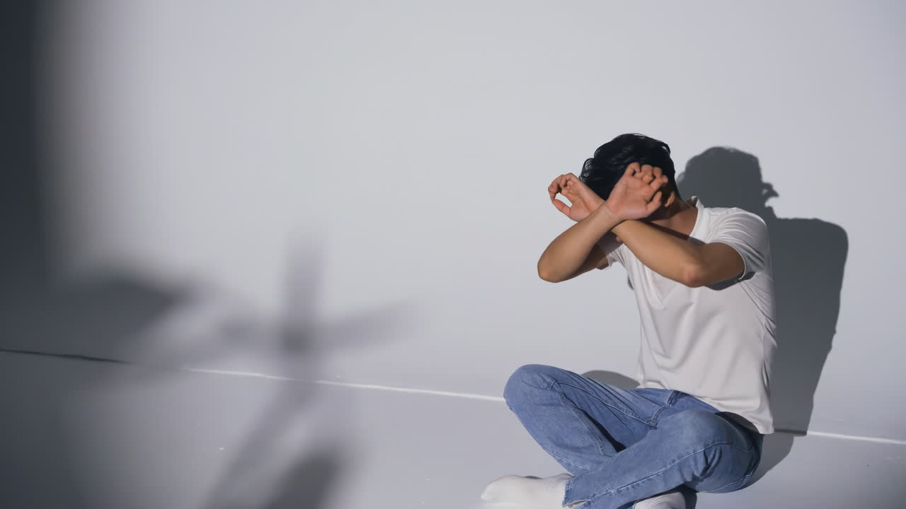 Un giovane uomo angosciato e stressato, che mostra paura e ansia per problemi di crisi nelle relazioni familiari. Si tiene la testa, mostrando dolore e angoscia a causa del conflitto, seduto per terra con una mano alzata in segno di stop.