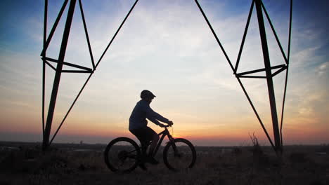 un ciclista de montaña masculino, se detiene debajo de una torre de línea eléctrica para observar el horizonte de una ciudad, mientras el sol bajo perfila su forma