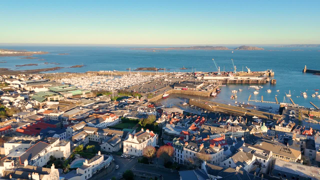High retreating drone footage of the roof tops of St Peter Port pulling back from harbour in the golden hour with views across to Herm and Jethou