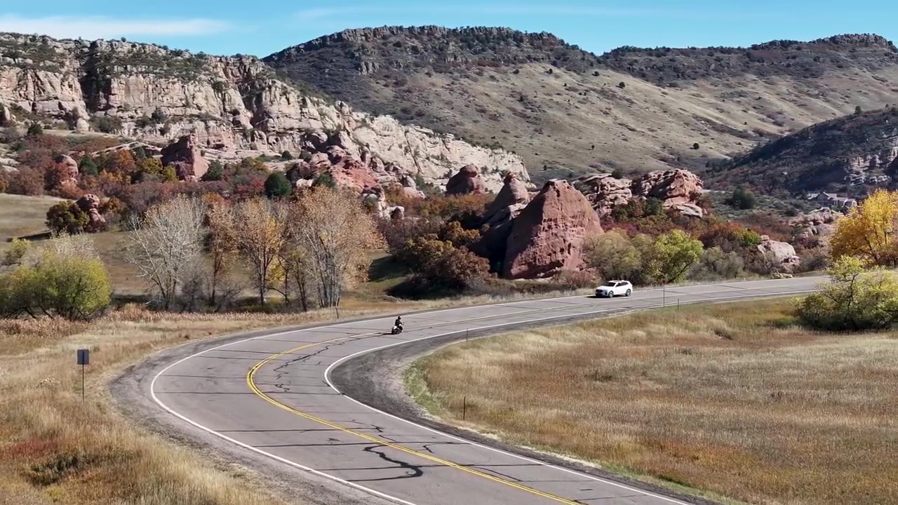 Toma cinematográfica aérea de dron de 70mm siguiendo a un motociclista solitario acelerando en una curva cerca del Cañón Deer Creek, rodeado de vívido follaje otoñal y llamativas formaciones rocosas rojas en Colorado.