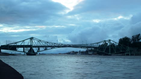 Lapso-De-Tiempo-De-Hermosas-Nubes-De-Lluvia-Gris-Oscuro-En-Movimiento-Rápido-Sobre-El-Puente-Oscilante-Oskara-Kalpaka-En-La-Ciudad-De-Liepaja-En-La-Noche,-Plano-General