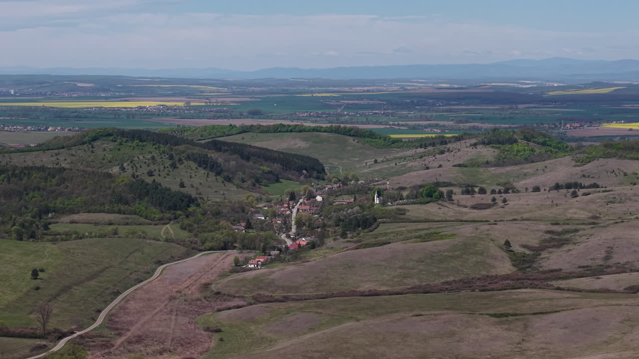 Un'inquadratura dall'alto cattura un tranquillo villaggio rurale situato in una valle verde, circondato da dolci colline e campi agricoli sotto un cielo sereno.