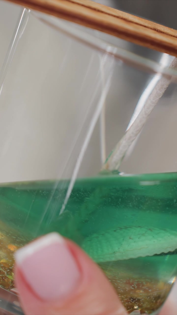 Primo piano di una mano che tiene un bicchiere con liquido verde e un filo bianco immerso. Il livello del liquido cambia man mano che la scena procede, mostrando l'interazione tra la texture dell'unghia e il vetro trasparente.