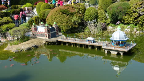 extreme wide shot of miniature promenade: orchestra performing in bandstand pavilion on tranquil green lagoon with reflections, serene waters, and intricate scenic details in a peaceful setting