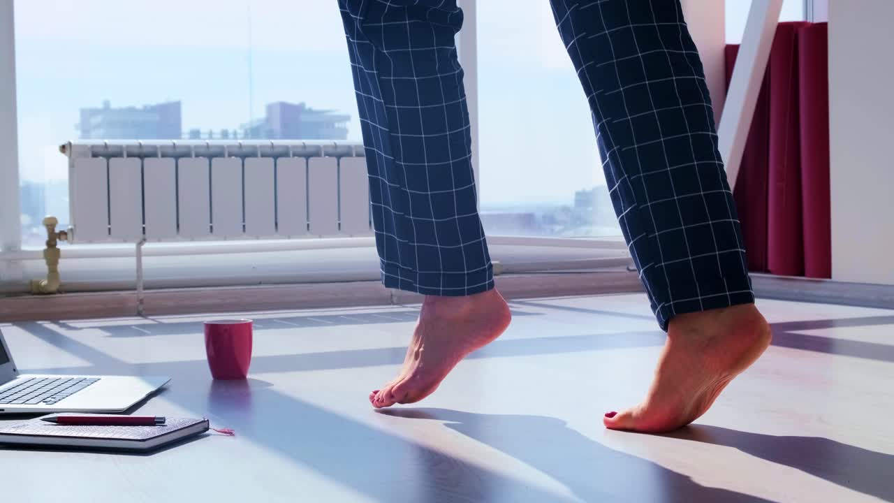 Morning Routine in a Sunlit Room: A Barefoot Individual Balances on Toes While Surrounded by a Laptop, Notebook, and Coffee in a Cozy Atmosphere