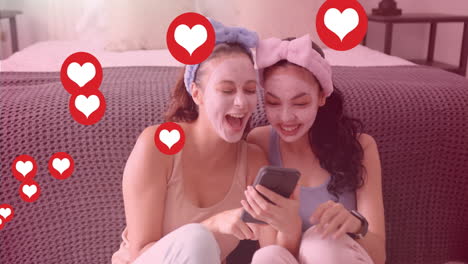 heart icons animation over two women with face masks laughing and using smartphone