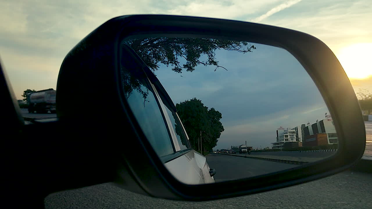 Specchietto retrovisore dell'auto che riflette il traffico autostradale e la strada sotto un cielo serale nuvoloso in India, mostrando i concetti di viaggio, tragitto, trasporto, movimento e stile di vita urbano.