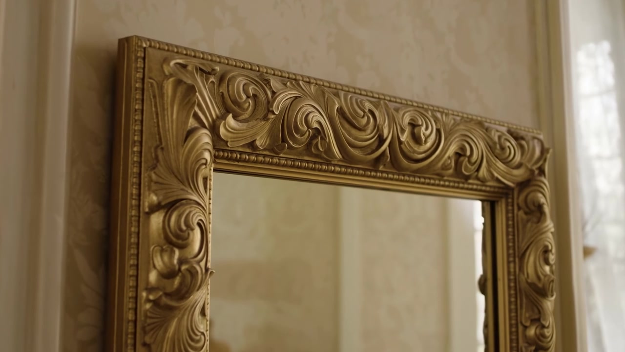 Close-up of an ornate gilded frame mounted on a beige wall, lit by soft daylight from a window at the right. The reflective glass and carved details create a