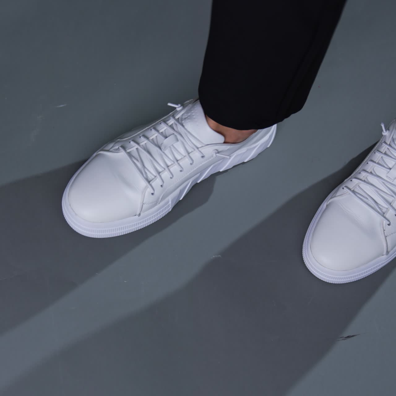 Modern snow-white sneakers on model's feet. Man in sport footwear taps his foot by the grey floor. Top view