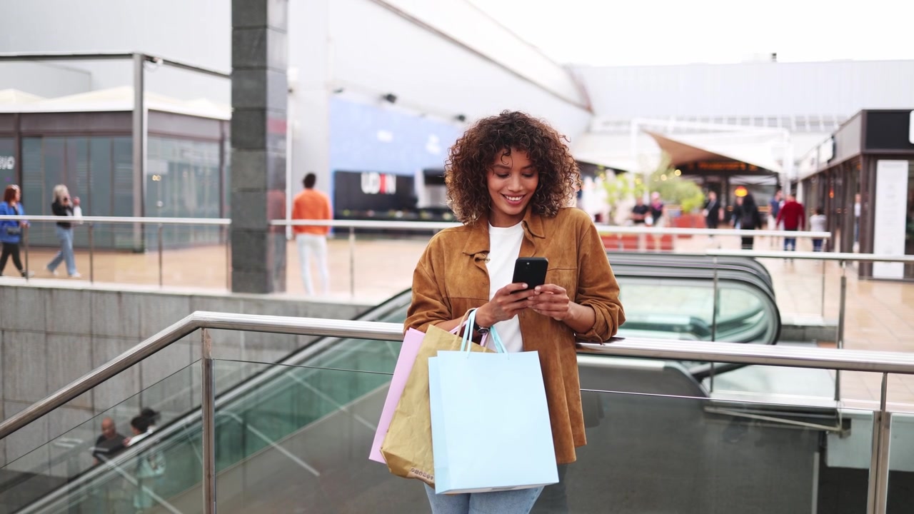 Happy Young Woman with Shopping Bags Using Phone in Mall