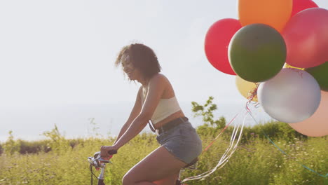 young woman riding bicycle decorated with balloons through countryside against flaring sun