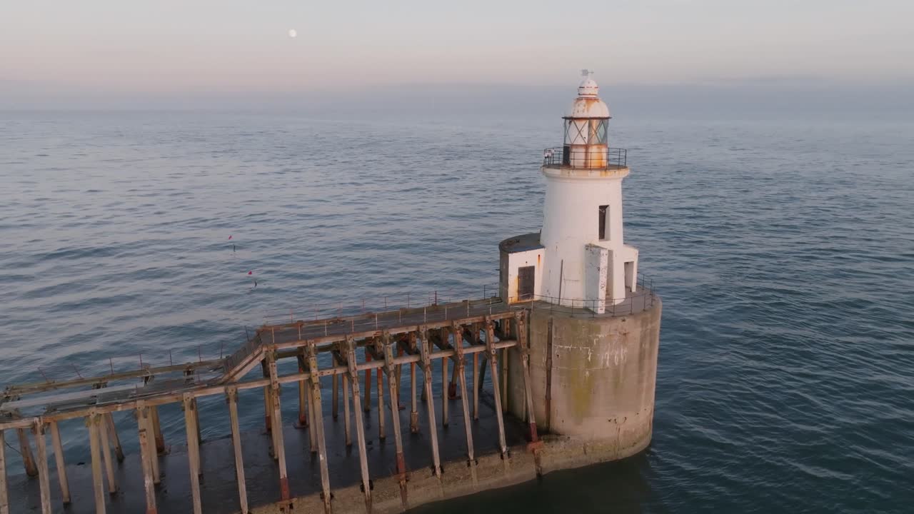 Traditional white lighthouse at end of harbour wall, at golden hour, with full moon and pastel skies
