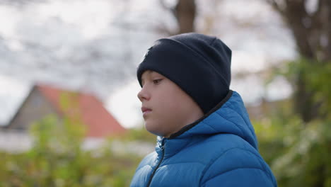 close-up of a young boy wearing a blue jacket and black beanie walking up to a tree, plucking leaves, with a blurred background featuring a distant building and vibrant greenery