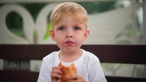 a young boy sits quietly, savoring a sausage roll, his expression is calm and focused as he slowly enjoys his snack