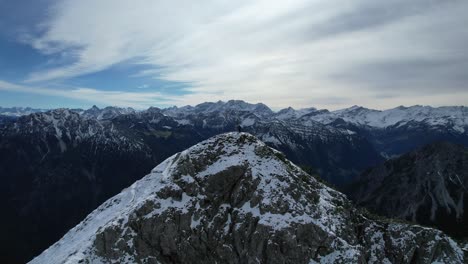 drone orbit shot of summit cross on snowy mountain peak of kuegrat during sunny day in liechtenstein. snowy peaks of mountains and green valley.