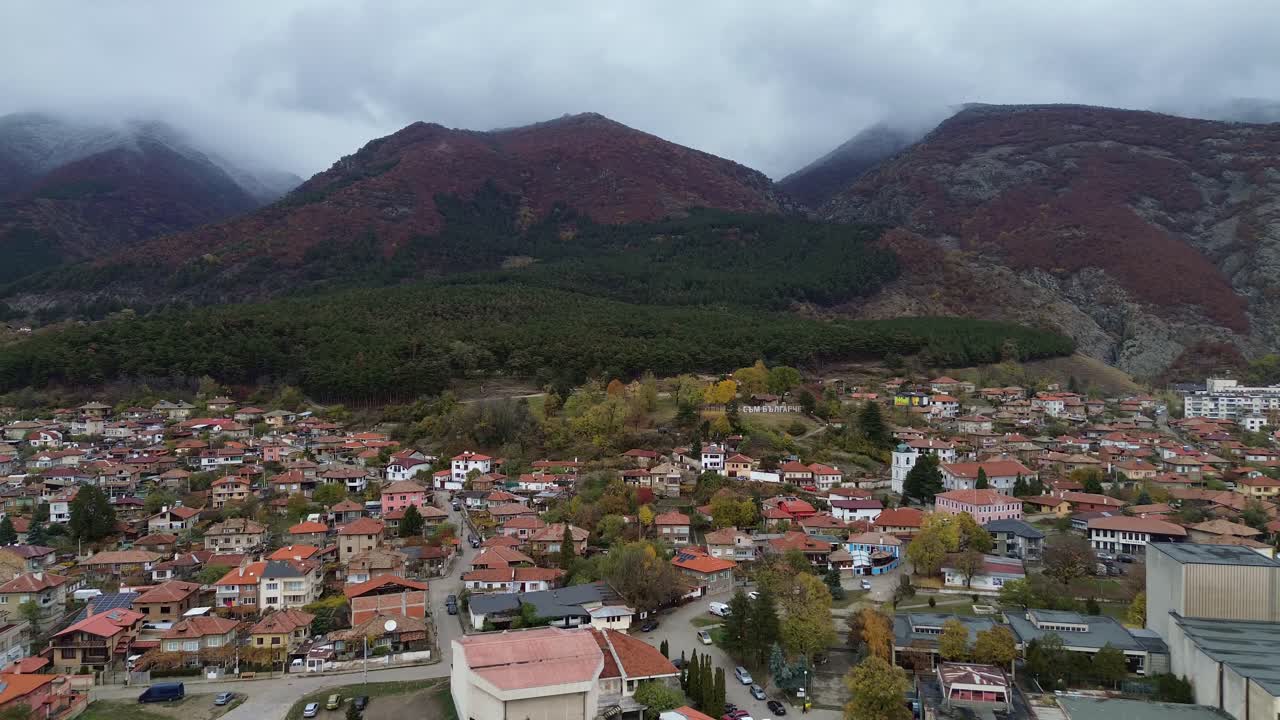 Drone footage over Sopot, Bulgaria, on a crisp winter day. The camera glides towards the Balkan Mountains, with the snow-covered peaks emerging beneath a soft, cloudy sky