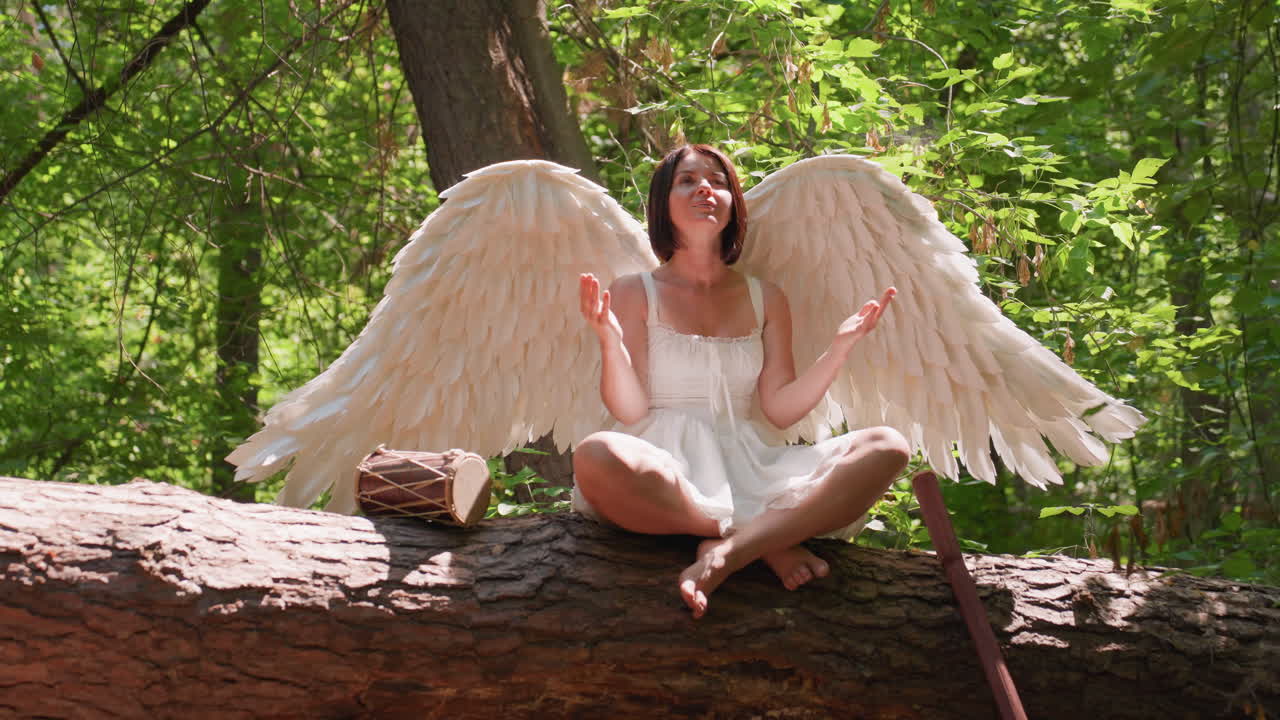 Actress with angelic wings dressed in white meditates to gods on fallen forest log surrounded by green leaves, talking drum and wooden staff nearby, sunlight casting warm glow