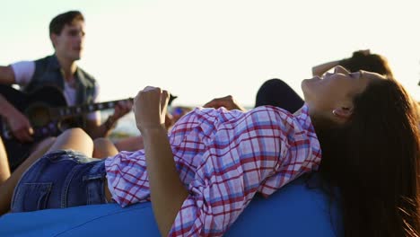 mujer joven acostada en una silla y escuchando a un hombre tocando la guitarra entre un grupo de amigos sentados en la playa y cantando en una noche de verano durante una puesta de sol. disparo en cámara lenta