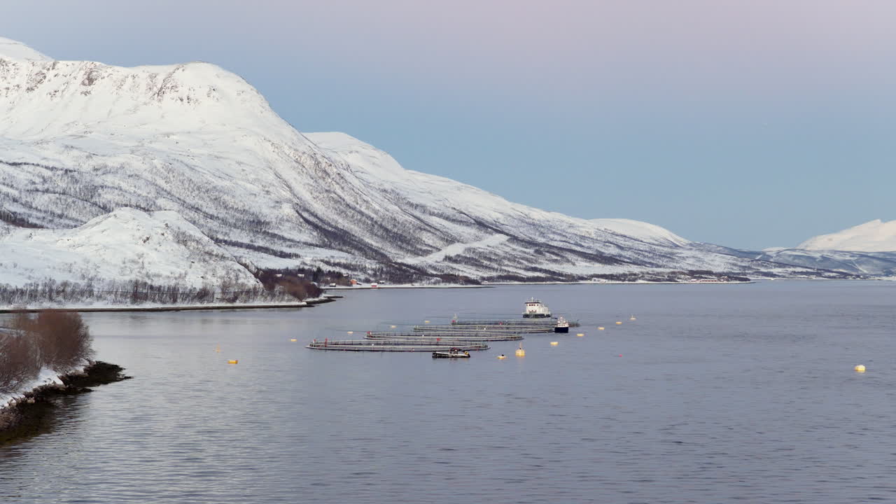 Vue aérienne d'une ferme aquacole de saumon avec un bateau de service, entourée de montagnes enneigées dans un fjord à Ringvassøya, Norvège