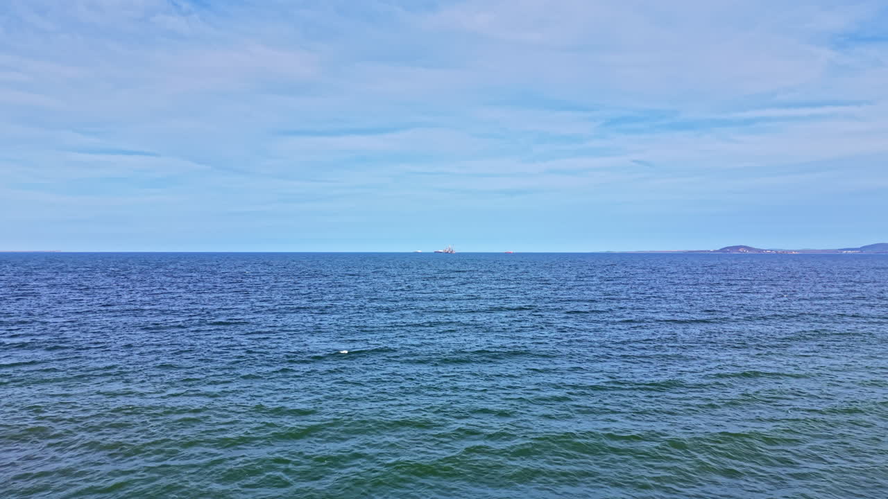 Bølger ruller på havet med en fjern landmasse under en blå himmel om dagen ved kysten.