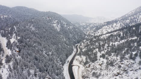 Aerial-reveal-forward-of-Boulder-Canyon-Drive-in-Colorado-during-the-winter-as-cars-drive-down-icy-road-surrounded-by-rocky-mountains-and-snow-covered-pine-trees