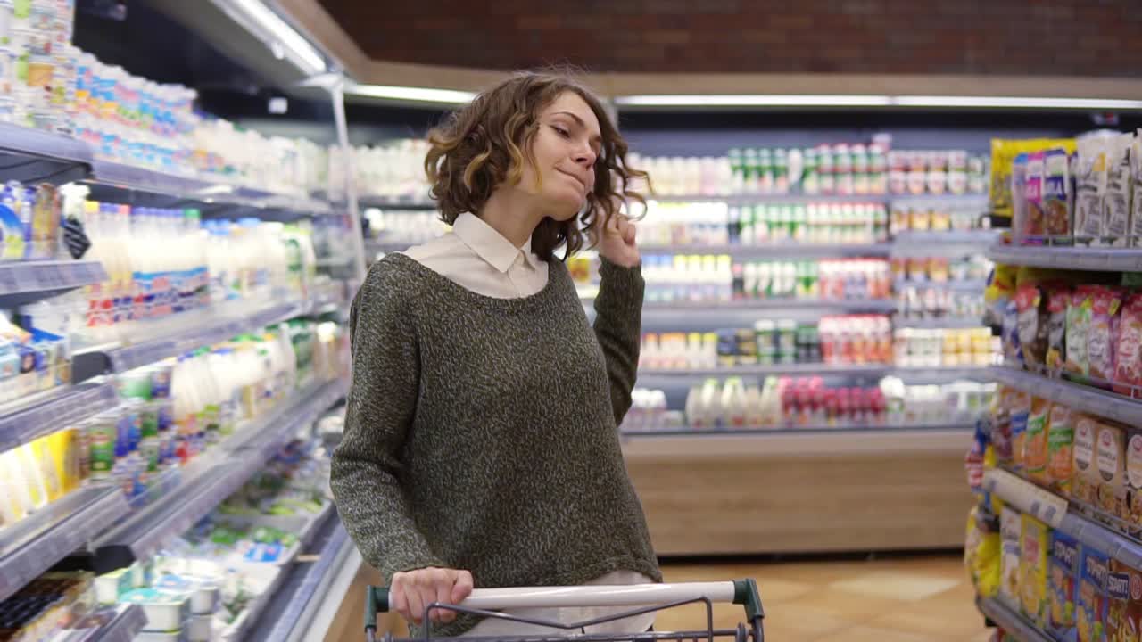 슈퍼마켓에서 행복한 젊은 여성은 선반에 있는 상품과 유제품을 통해 춤을 추고 있습니다. 회전하고, 재미있고, 긍정적입니다. 쇼핑 카트를 밀고 있습니다. 전면 전망