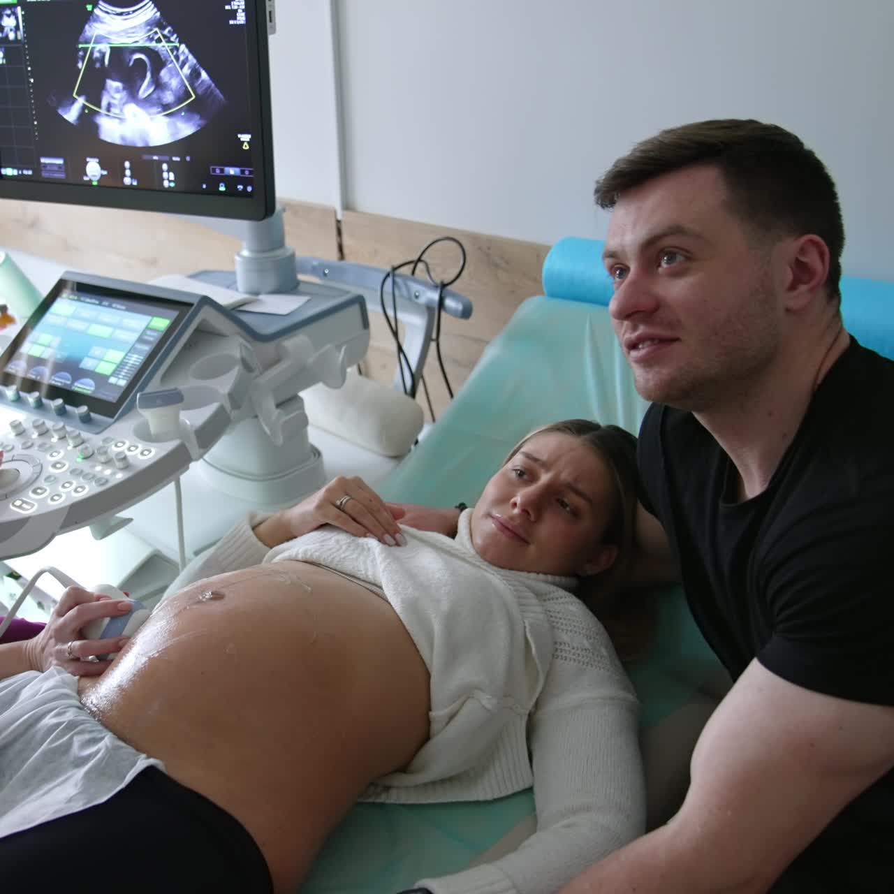 Expecting family are having ultrasonic check up at obstetrician's cabinet. Man supports his wife lying on a couch. Happy parents looking a baby image on the screen and smile happily