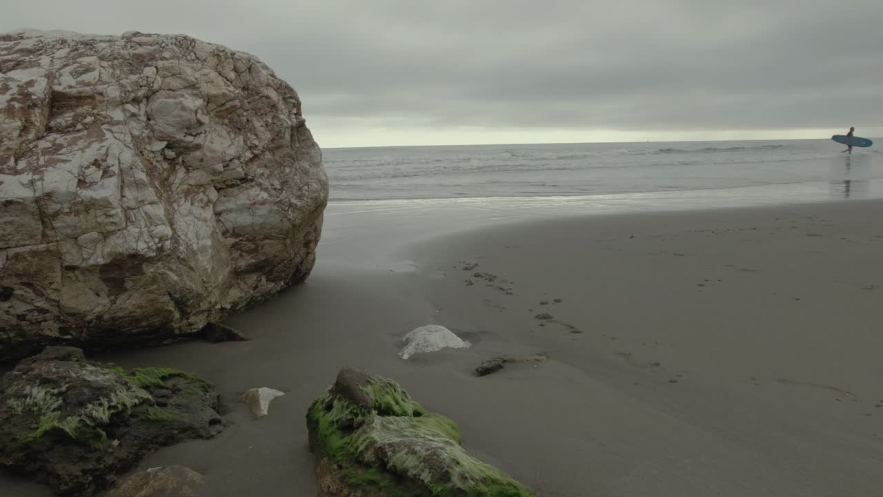 Detail of a beach with a huge rock while a young surfer gets out of the sea walking along the shore.