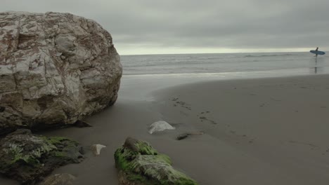 detail of a beach with a huge rock while a young surfer gets out of the sea walking along the shore.